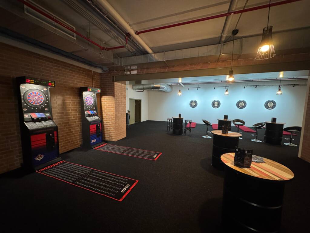 Ein großer Dart-Raum mit zwei elektronischen Dartautomaten links und vier klassischen Dartboards an der entfernten Wand. Dazwischen stehen mehrere hohe Fass-Tische mit Barhockern in gedimmter Beleuchtung.