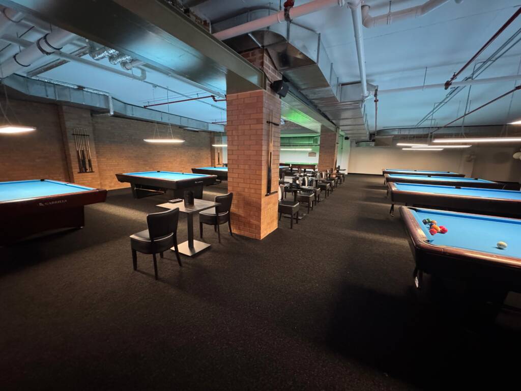 Ein Poolbillardraum mit mehreren blau beleuchteten Tischen, Sitzbereichen zwischen den Tischen und sichtbarer Deckeninstallation in einer weitläufigen Halle.