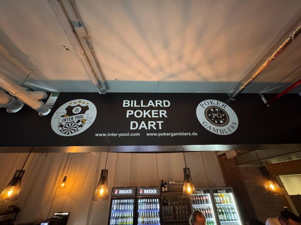 Ein Schild über einer Bar zeigt die Wörter „Billard, Poker, Dart“ sowie zwei Logos und Webseiten, darunter Getränke-Kühlschränke und warm beleuchtete Hängelampen.