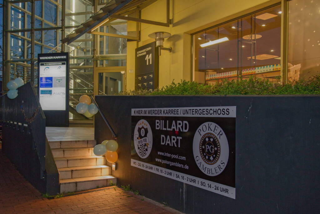 Außenansicht eines Gebäudeeingangs bei Abendlicht, mit Treppe, Luftballons und einem großen Werbeschild für Billard, Dart und Poker im Untergeschoss.