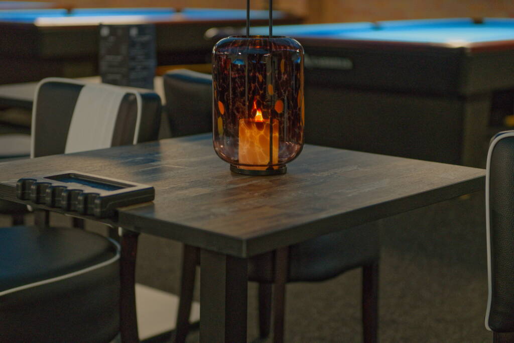 Ein kleiner Holztisch mit einem dunklen Glaswindlicht und elektrischer Kerze, daneben ein Bediengerät; im Hintergrund unscharfe Billardtische.