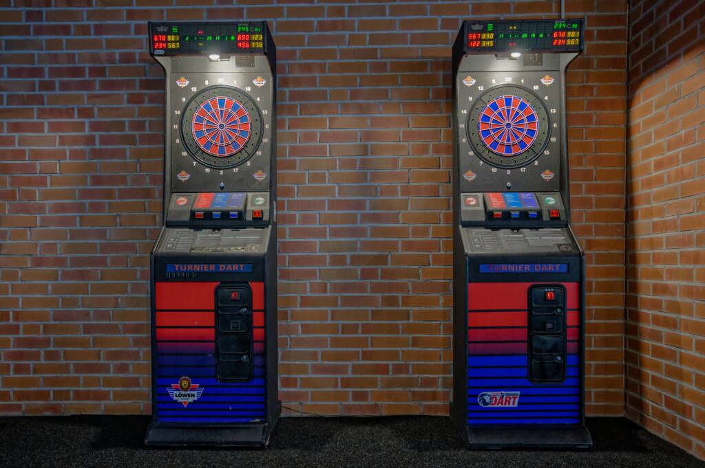 Zwei elektronische Turnier-Dartautomaten stehen nebeneinander vor einer Backsteinwand, jeweils mit beleuchteter Dartscheibe und digitalem Punktedisplay.