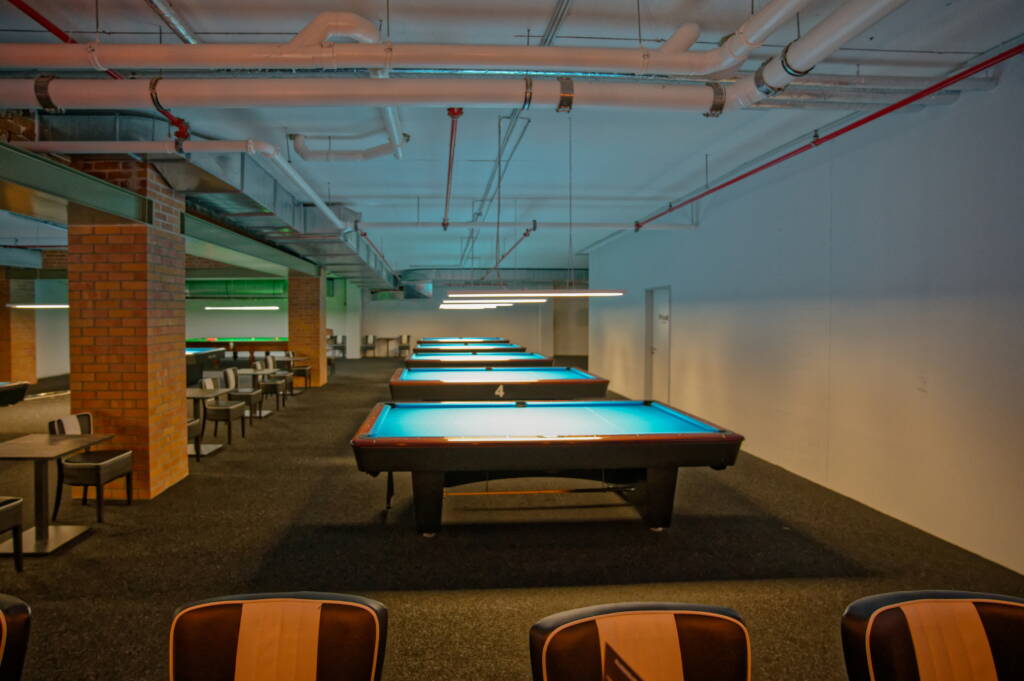 Mehrere blau beleuchtete Pooltische stehen in einer Reihe unter Deckenleuchten, links daneben Sitzbereiche mit Tischen und Stühlen in einem weitläufigen Raum.