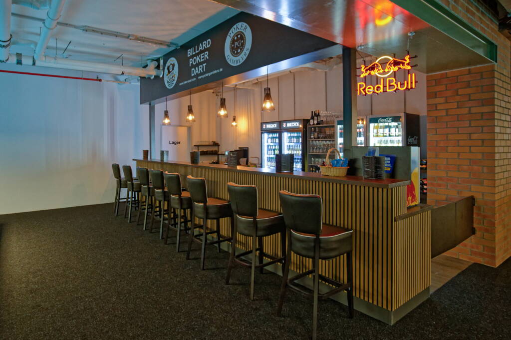 Eine lange Bar mit hohen Stühlen, beleuchteten Getränkekühlschränken und einem Red-Bull-Neonschild; darüber ein Schild mit „Billard Poker Dart“.