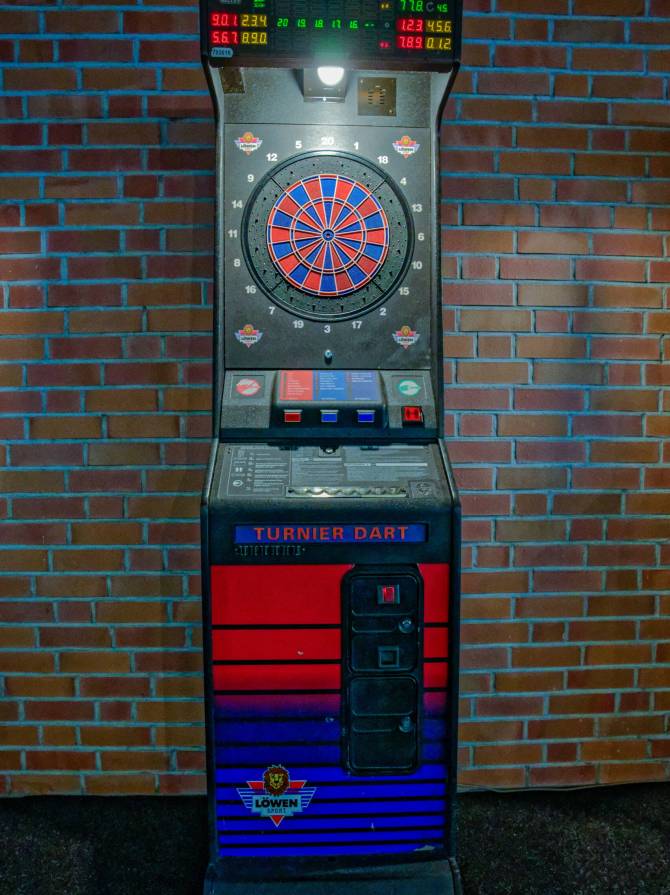 Ein elektronischer Turnier-Dartautomat vor einer Backsteinwand, mit beleuchteter Dartscheibe und digitalem Punktedisplay.