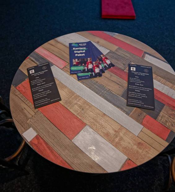 Ein runder Holztisch mit bunten Holzlatten-Muster, darauf mehrere Flyer und Visitenkarten; daneben stehen zwei Barhocker.