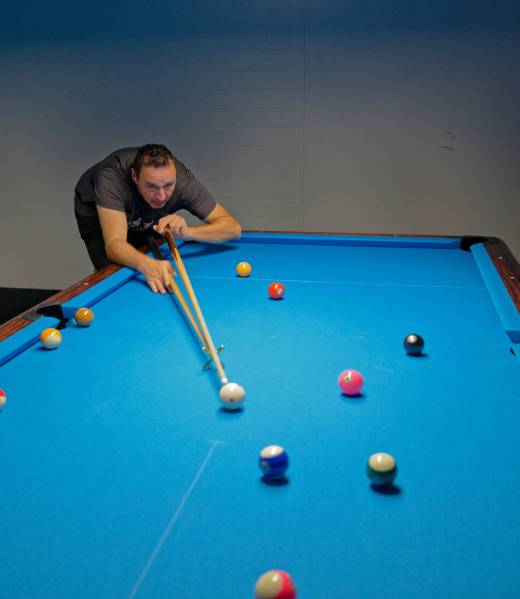 Ein Mann beugt sich über einen blau beleuchteten Pooltisch und stößt mithilfe einer Brücke die weiße Kugel an; mehrere bunte Kugeln liegen verteilt auf dem Tisch.