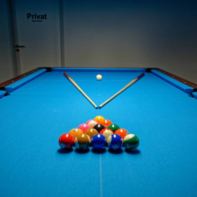 Ein Pooltisch mit aufgebautem Dreieck bunter Kugeln im Vordergrund, zwei Queues in V-Form dahinter und der weißen Kugel vor einer Tür mit der Aufschrift „Privat“.
