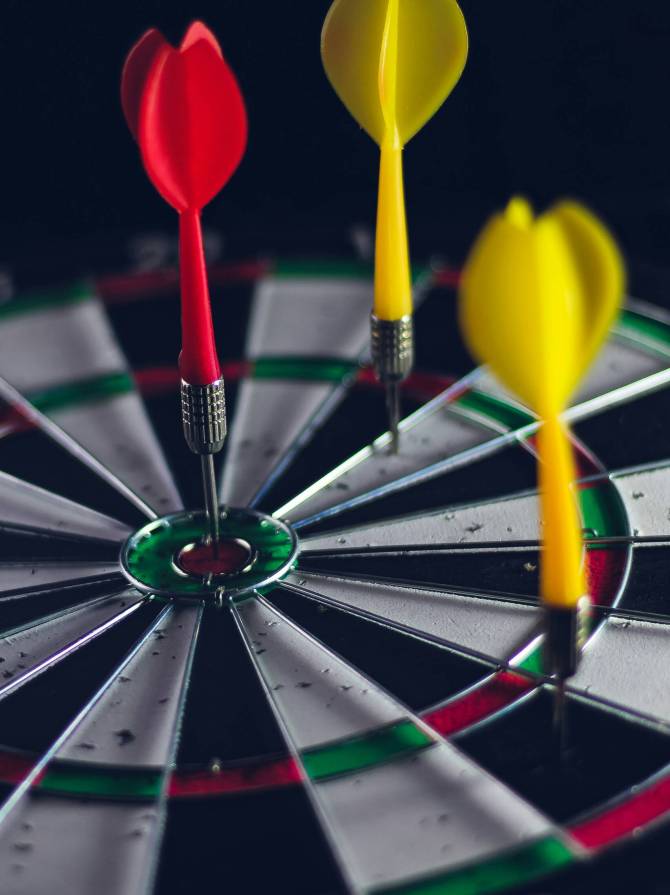 Drei Pfeile stecken in einer Dartscheibe, einer im roten Bullseye und zwei gelbe daneben.