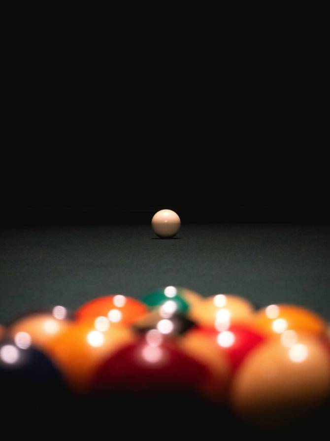 Eine hell beleuchtete weiße Spielkugel steht auf einem dunklen Pooltisch, im Vordergrund unscharf das bunte Dreieck der aufgebauten Kugeln.